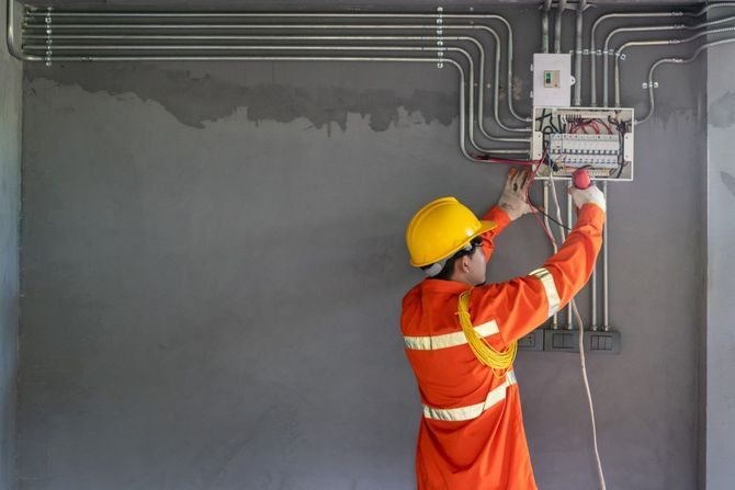 Worker in orange safety gear inspecting electrical wiring in a gray-walled room