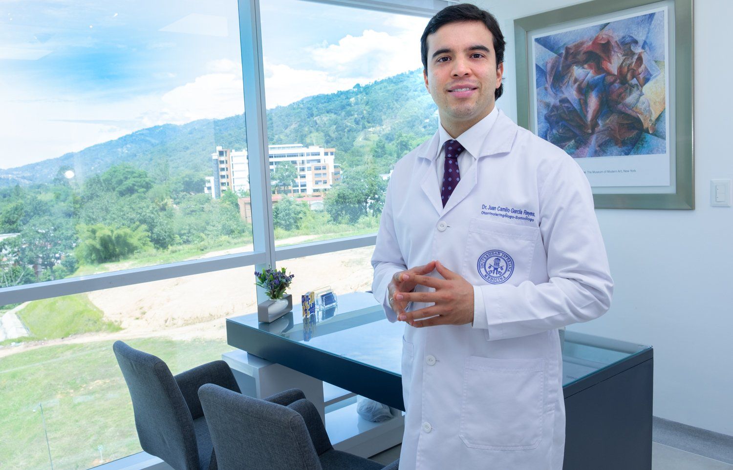 Un médico con bata blanca está de pie en su consultorio con ventana y vista a la montaña.