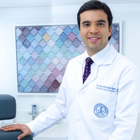 Médico sonriente con bata blanca, de pie cerca de una silla y un lavabo, con arte de pared con dibujos azules.