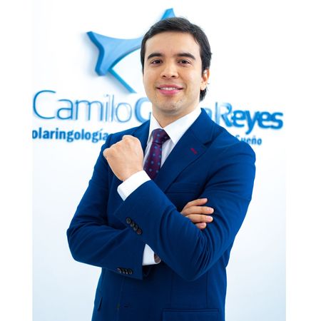 Hombre de traje azul, sonriente, con los brazos cruzados, frente al logo de Camilo Reyes, otorrinolaringólogo.