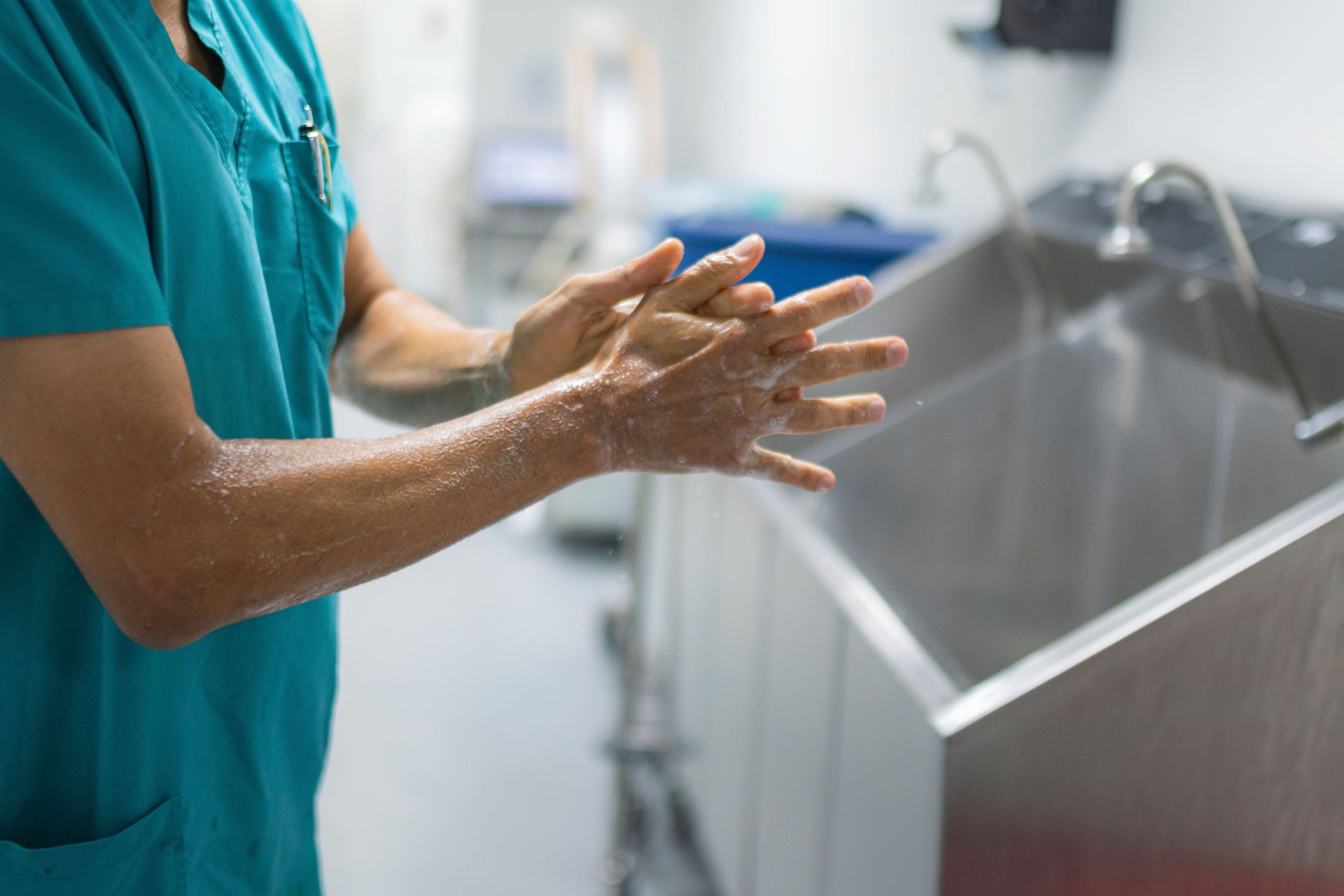 Pessoa vestindo uniforme cirúrgico verde-azulado lavando as mãos em uma pia de aço inoxidável em um ambiente médico.