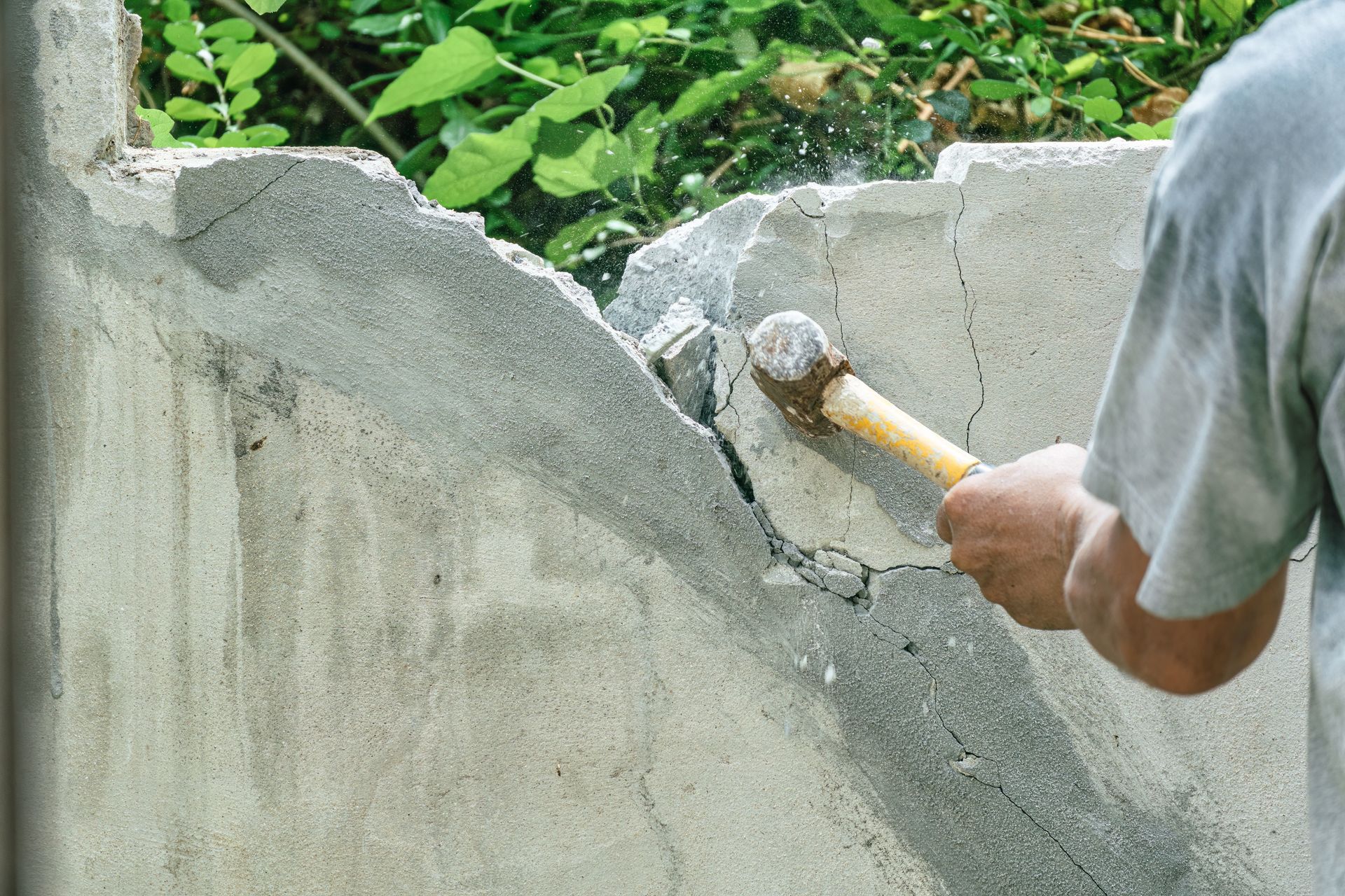 Person using a sledgehammer to break apart a concrete wall outdoors.