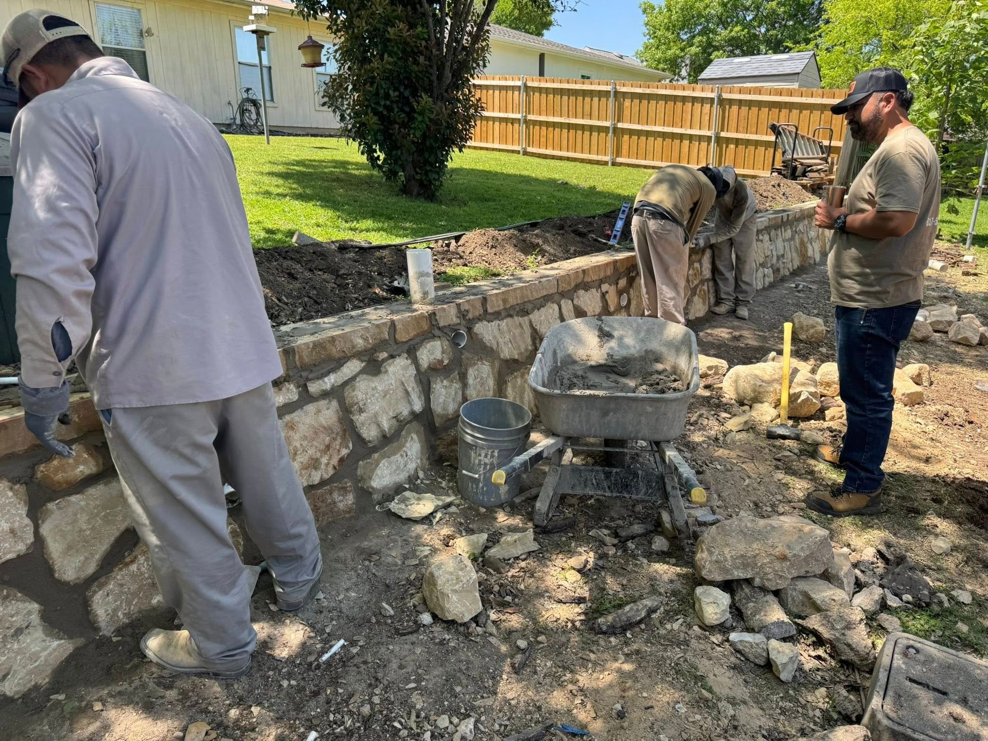 Construction workers building a stone retaining wall in a sunny backyard.