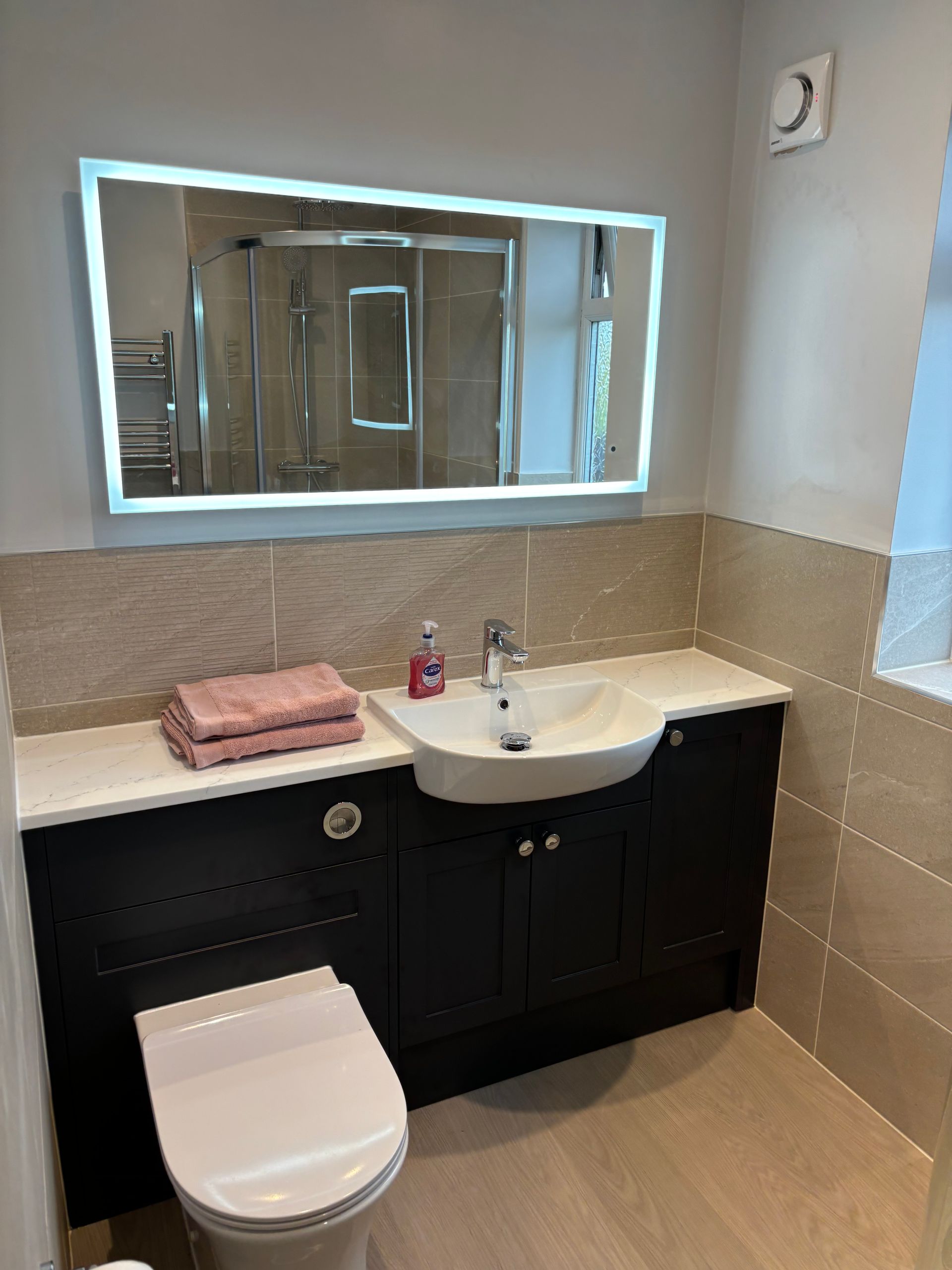 Modern bathroom with black vanity, illuminated mirror, sink, toilet, and beige tiled walls