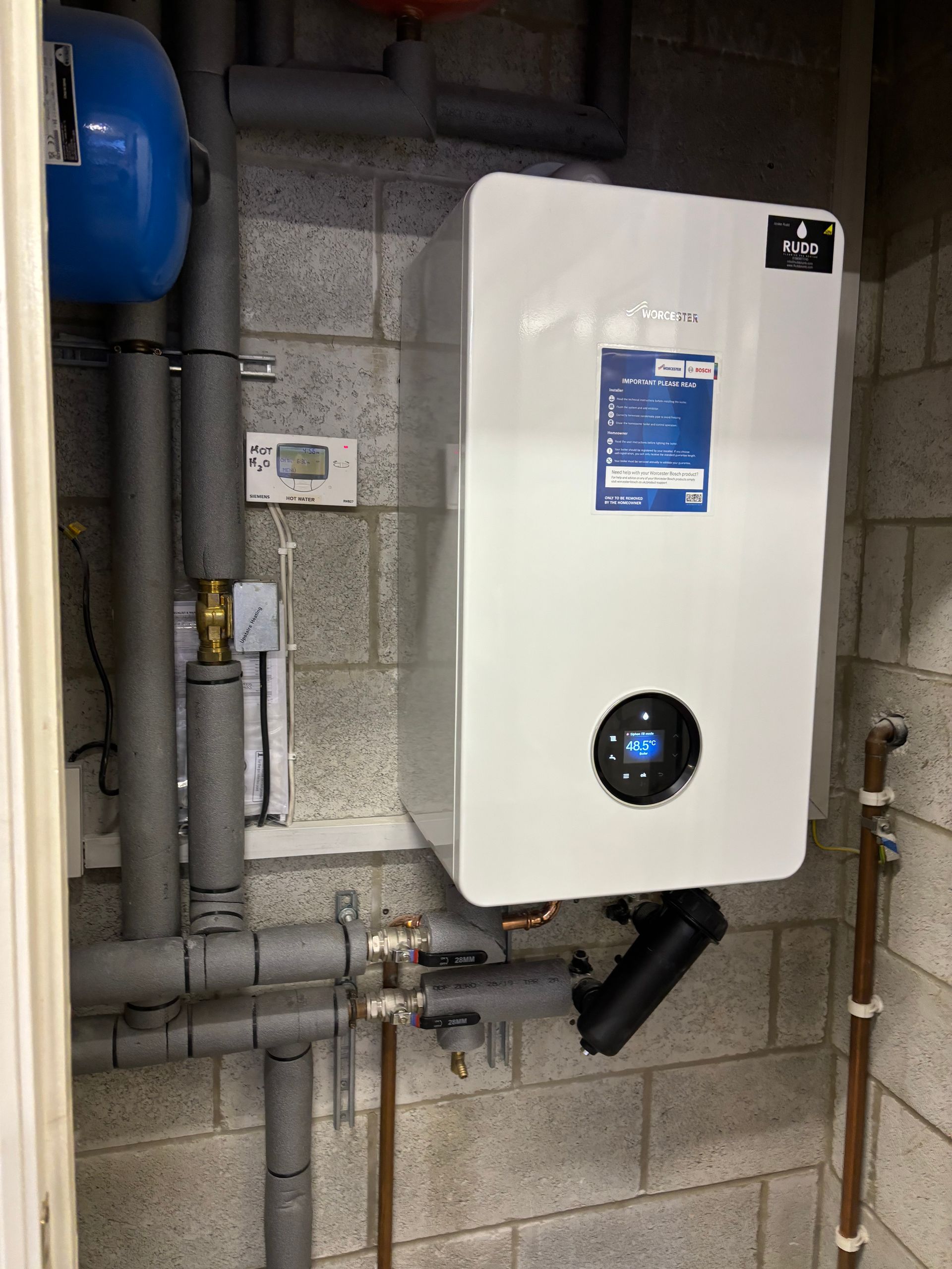 White wall-mounted boiler with blue display in a utility room, surrounded by exposed pipes and cinder block walls