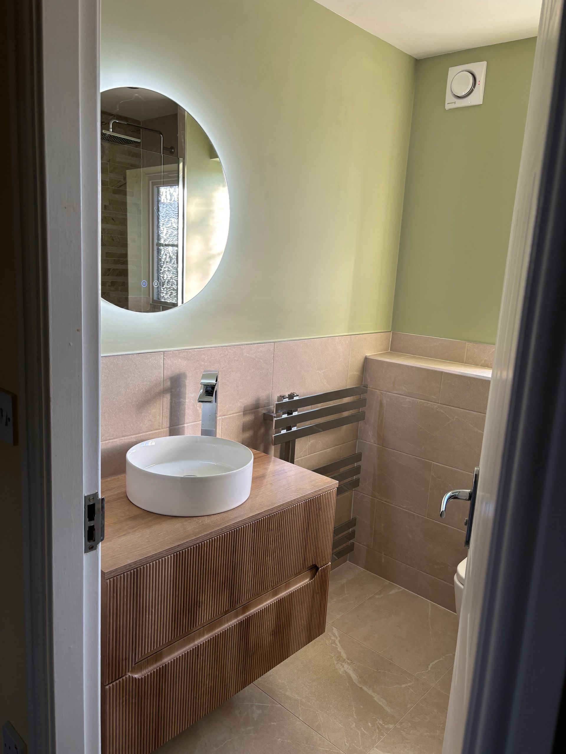 Small bathroom with round mirror, white vessel sink, beige tiles, and pastel green walls.