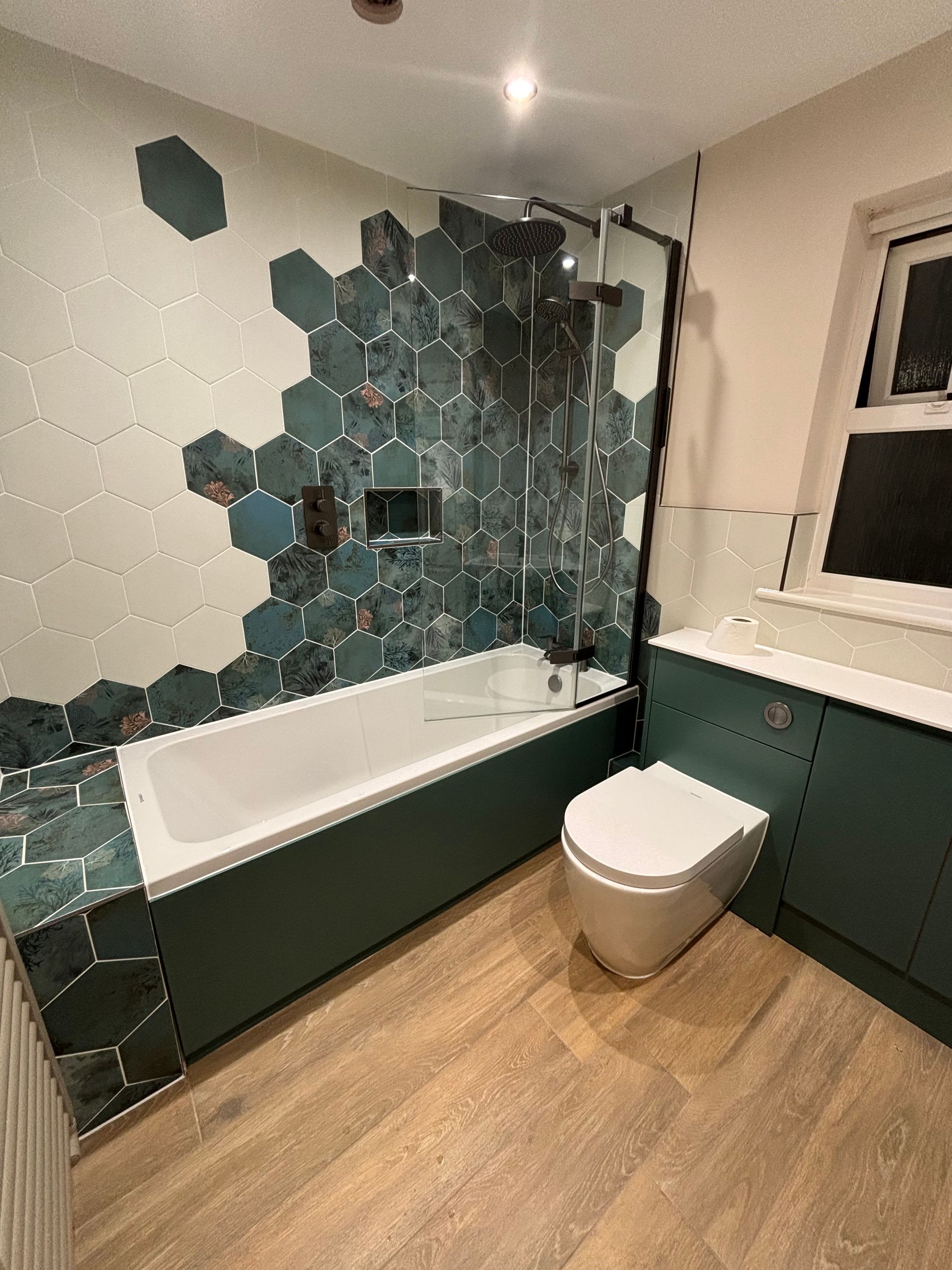 Modern bathroom with teal hex tile accent wall, white tub, and dark vanity beside a toilet