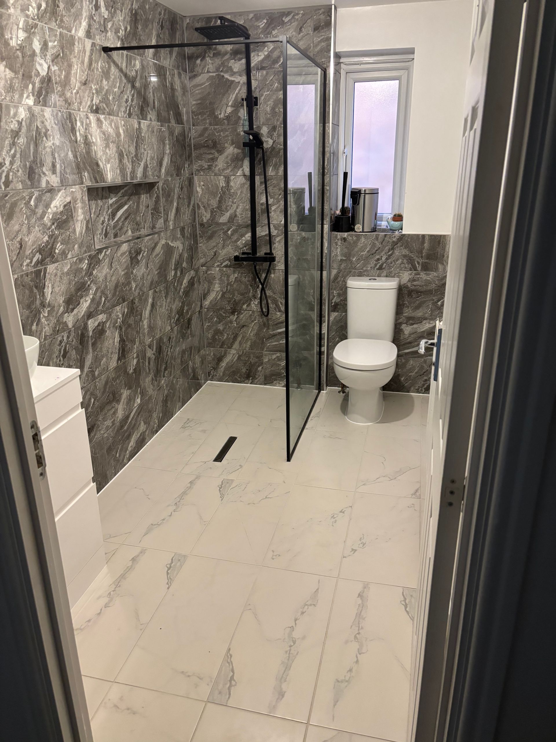 Small modern bathroom with marble walls, glass shower, toilet, and white tiled floor.