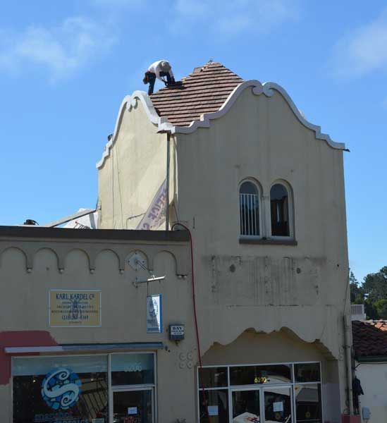 Two people on the roof of a light-colored building. The building has arched windows and a storefront. Blue sky.