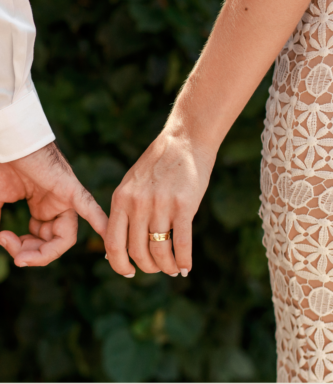 Two hands holding against a blurred green outdoor background, one wearing a gold ring
