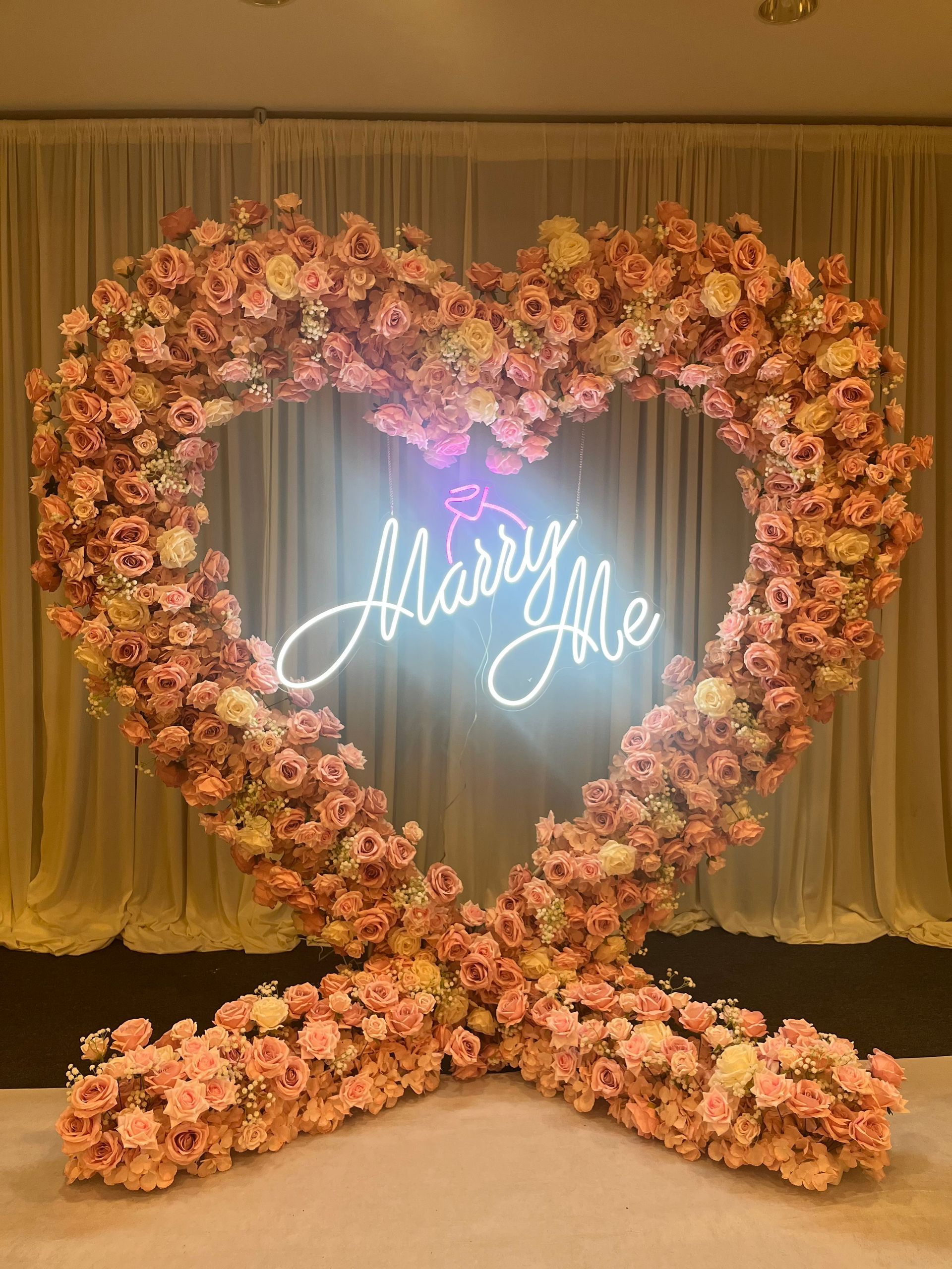 Heart-shaped floral arrangement with neon “Marry Me” sign against gold drapes