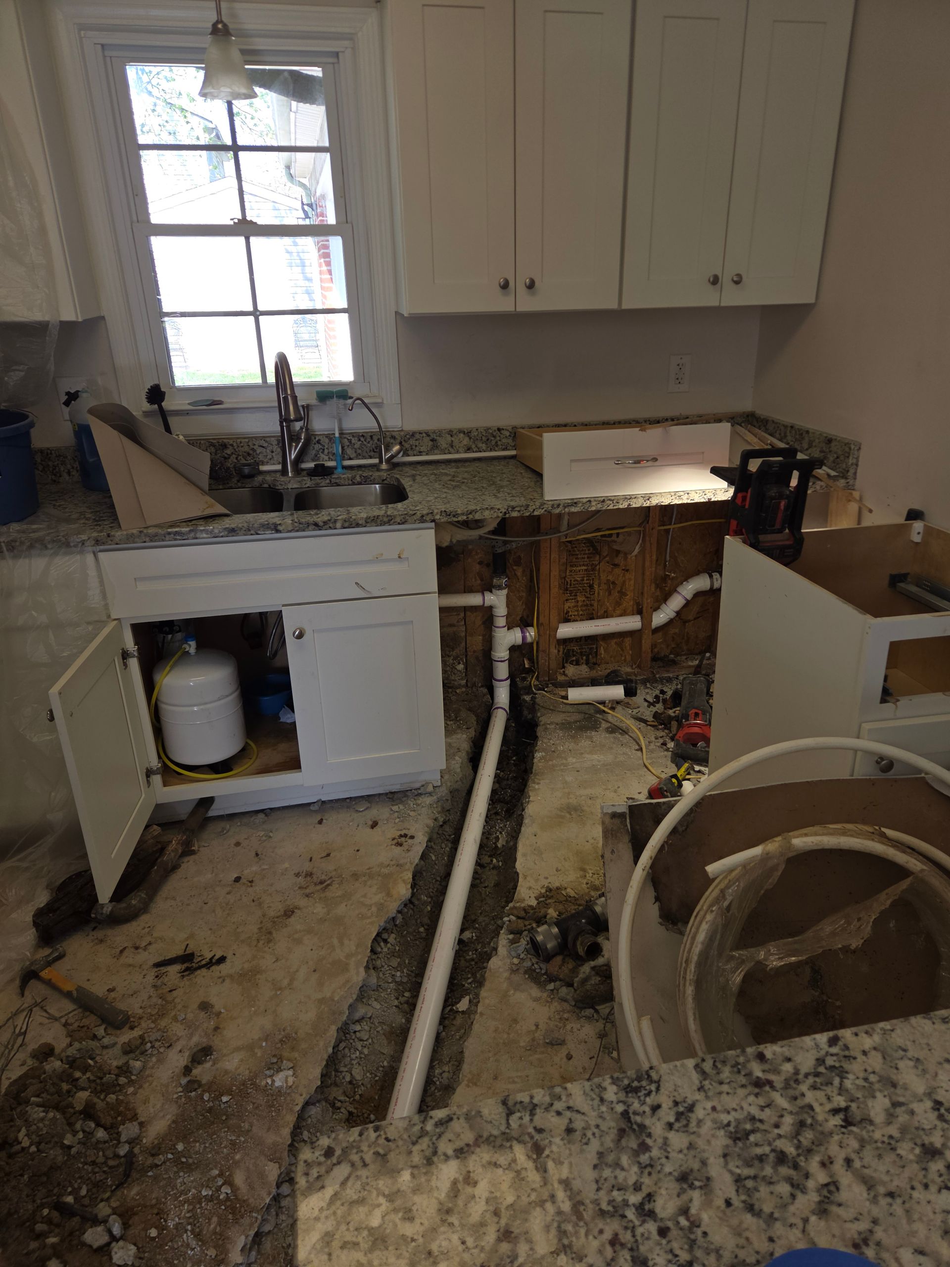 Damaged kitchen with torn-up flooring, exposed subfloor, open cabinets, and debris scattered around.