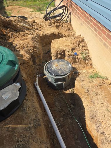 Backyard excavation around a round utility lid beside a brick house and green tank.