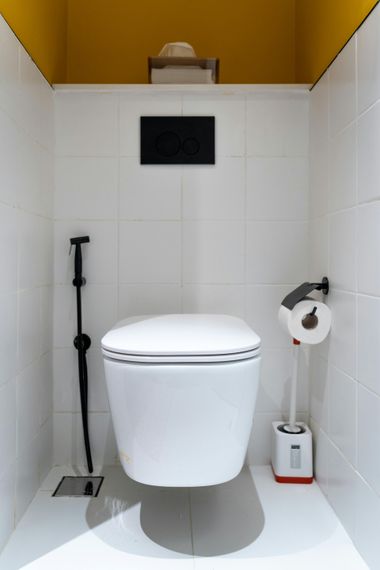 Minimalist white toilet stall with a wall-mounted toilet, black flush panel, and toilet paper holder.
