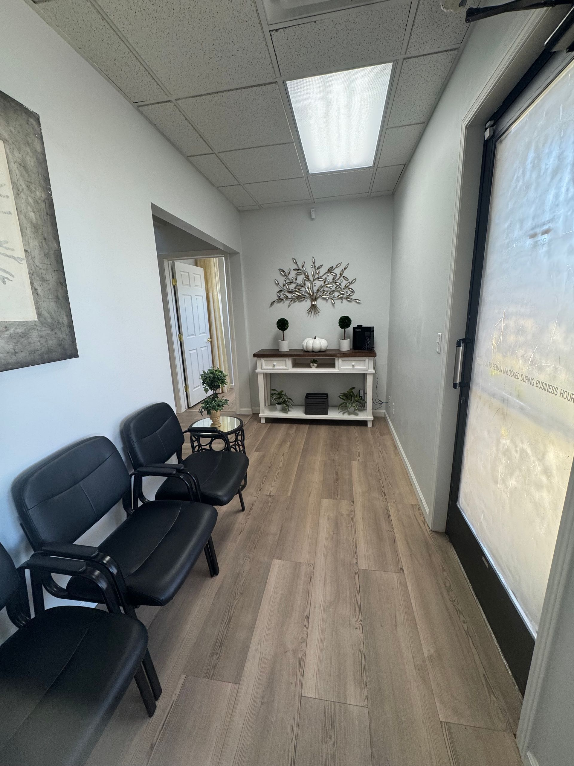 Narrow waiting room with black chairs, wood floor, white walls, and a reception table by a frosted window