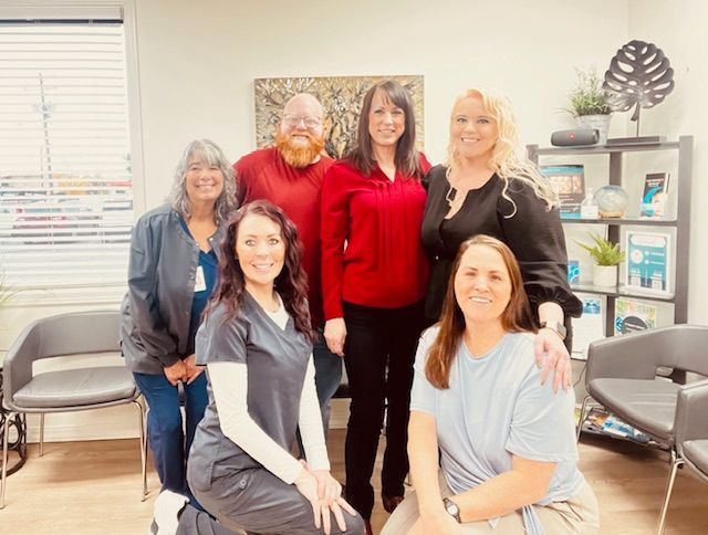 Six people posing in a bright office, with chairs, shelves, and a leaf wall decor in the background.