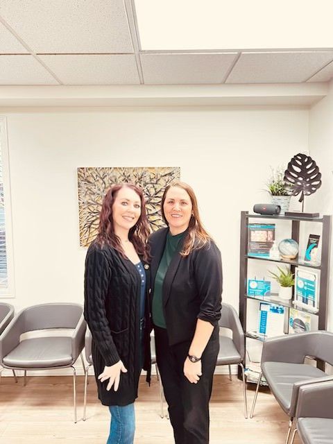 Two women standing and smiling in a bright office with chairs and a shelf of supplies.