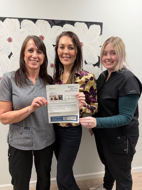 Three women stand indoors, smiling and holding a certificate in front of a heart-decorated wall.