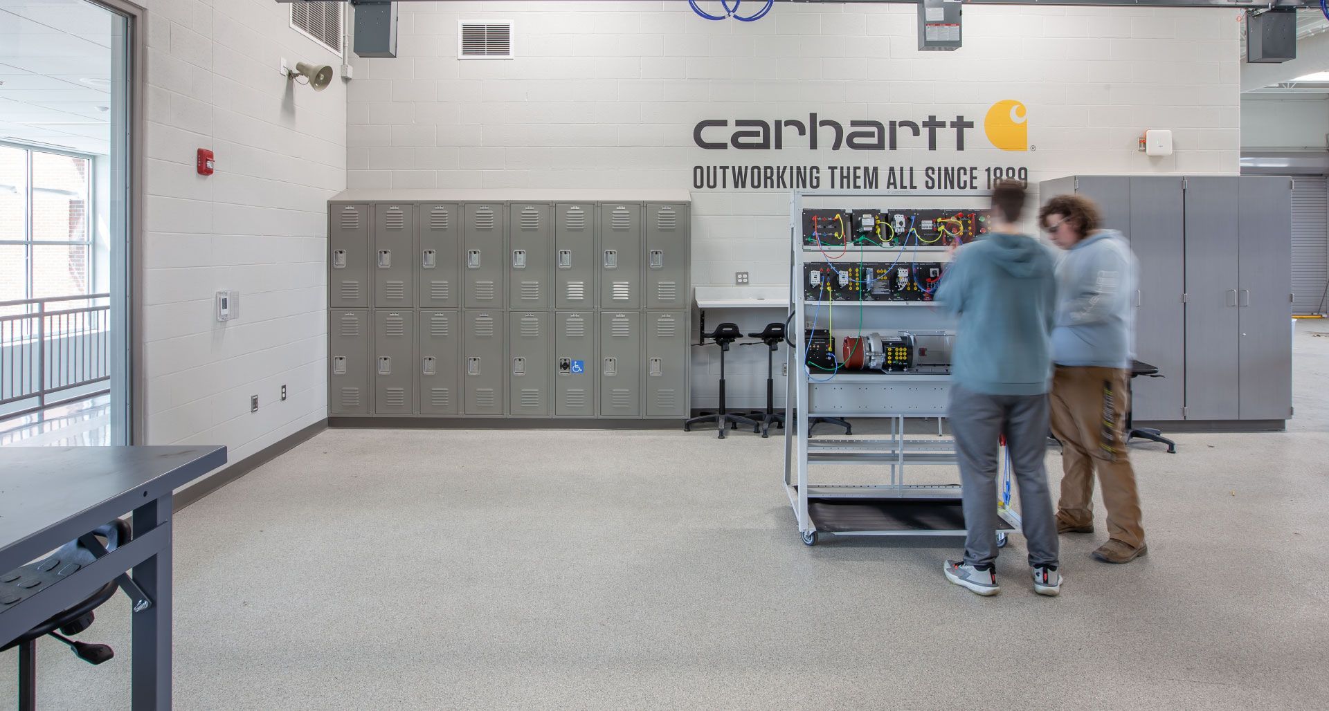 Two people are standing in a room with lockers and a carhartt logo on the wall.