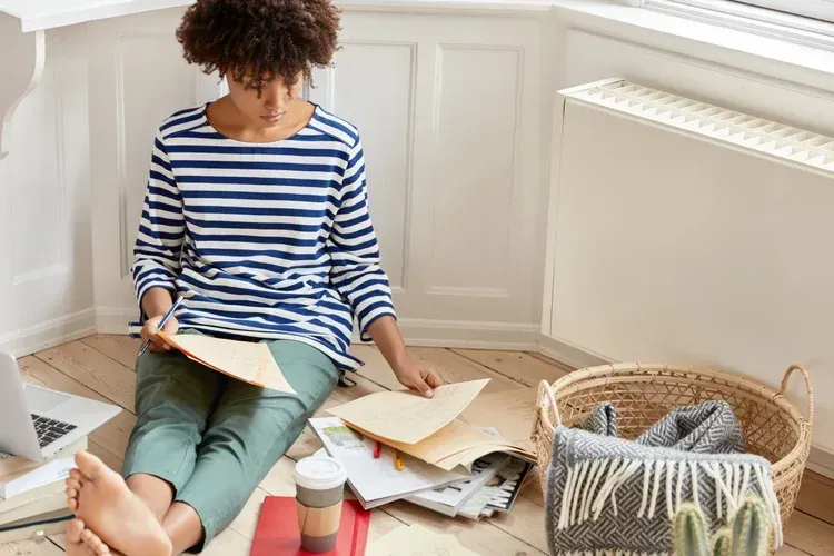 Person sitting on the floor reading books beside a laptop, notebook, and woven basket in a bright room