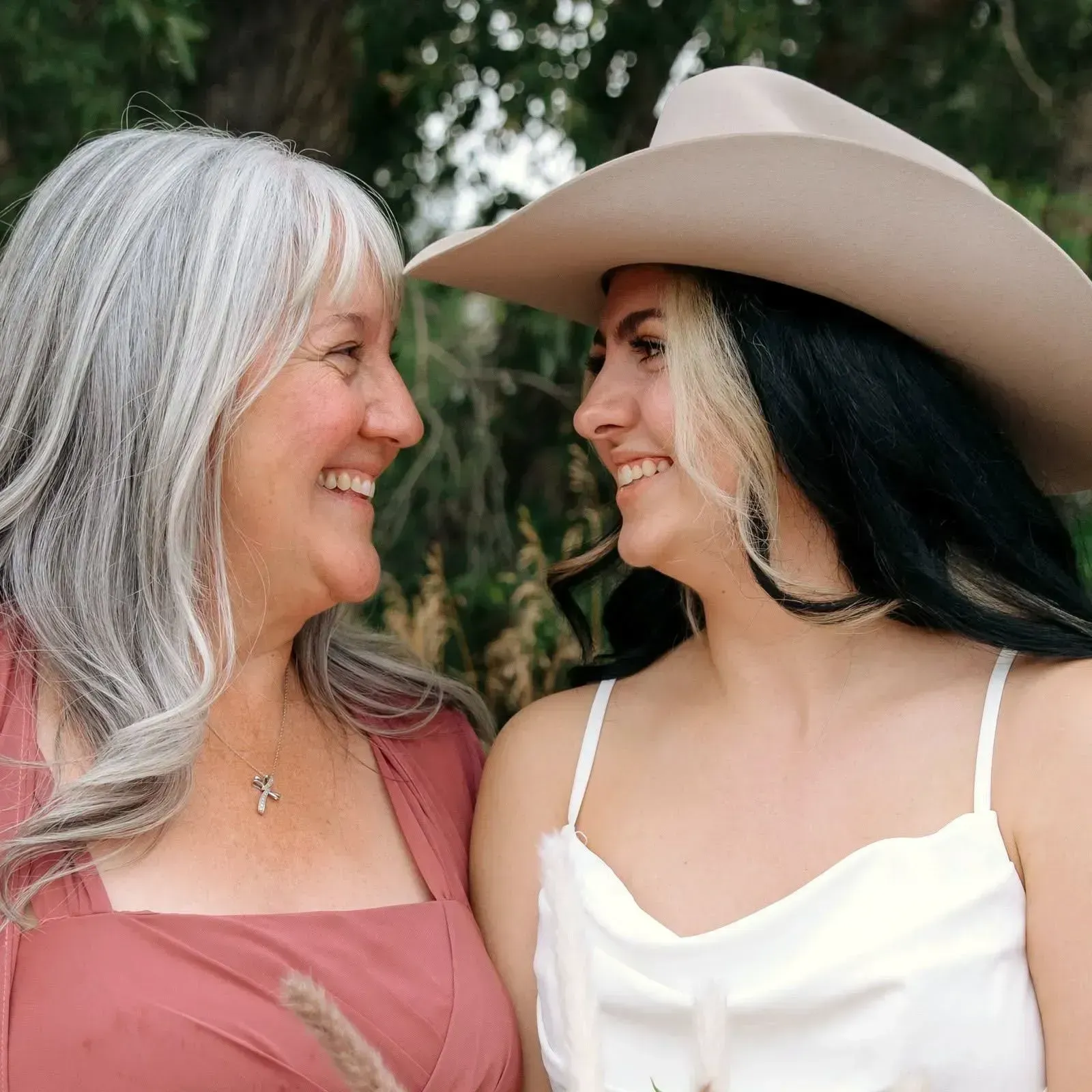 Two women smiling at each other outdoors, one in a cowboy hat and white top, the other in a pink dress.