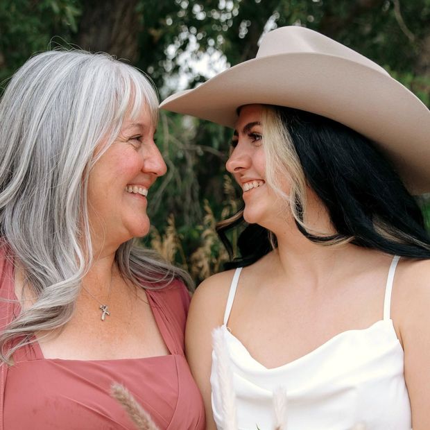 Two women smiling at each other outdoors, one in a cowboy hat and white top, the other in a pink dress.