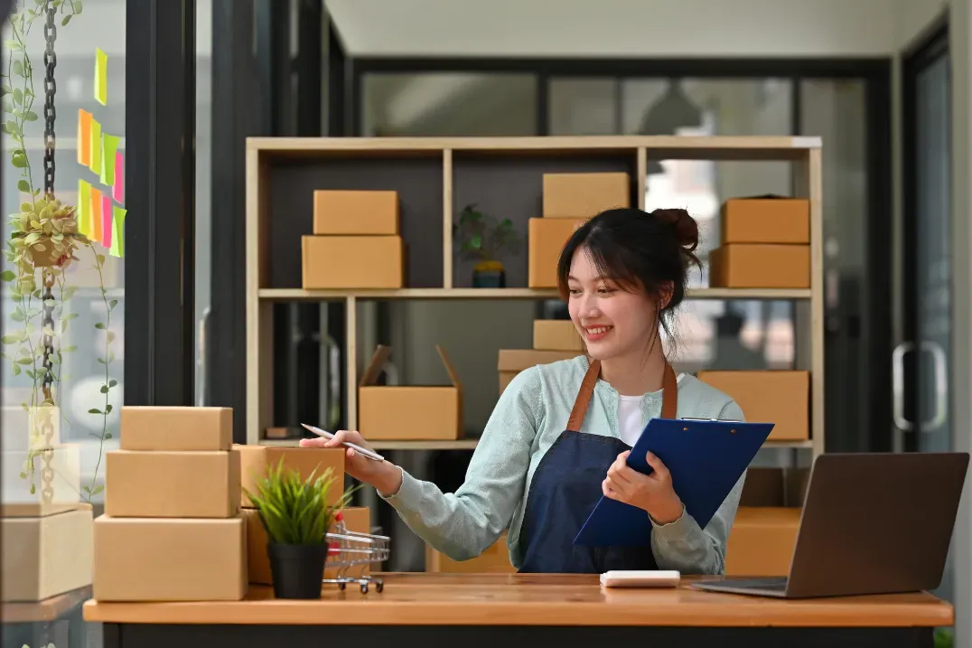 Person packing boxes at a desk in a warehouse, holding a clipboard beside shelves and a laptop