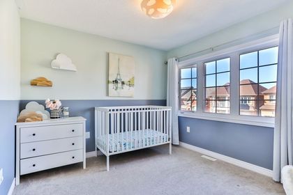 Bright nursery with a white crib, dresser, cloud wall decor, and large window overlooking houses.