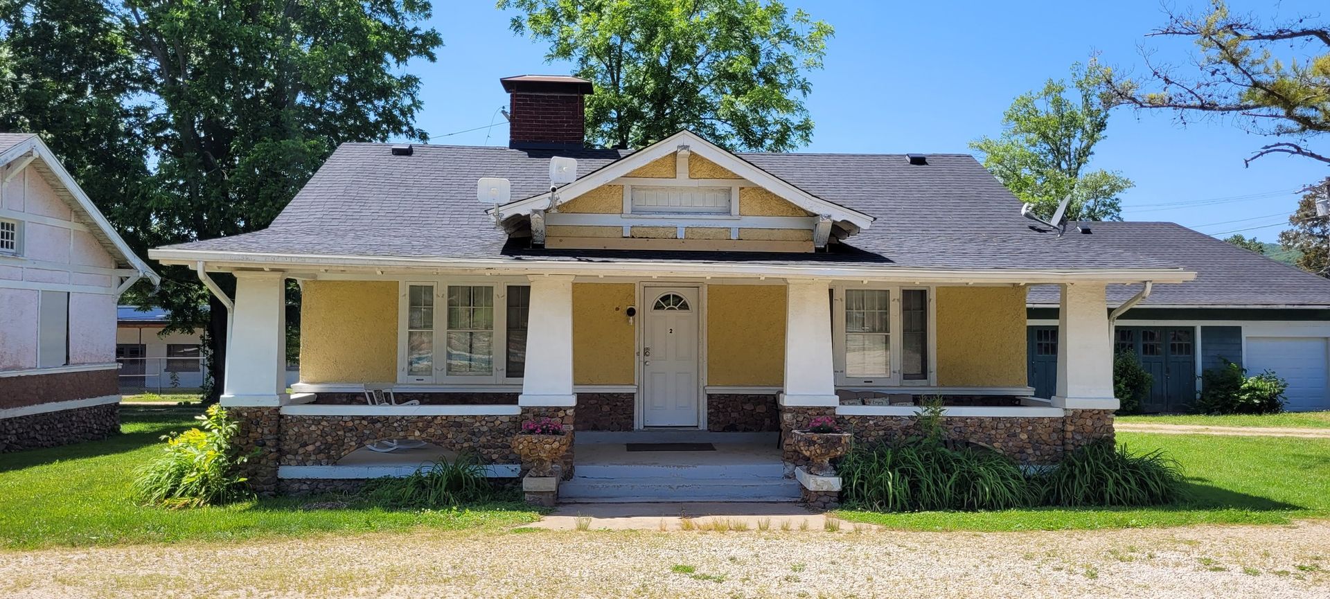 A yellow house with a porch and a gray roof is sitting on top of a lush green field.