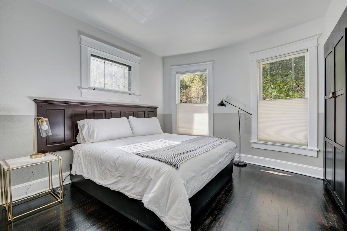 Bright bedroom with dark wood bed, white bedding, and large windows with blinds