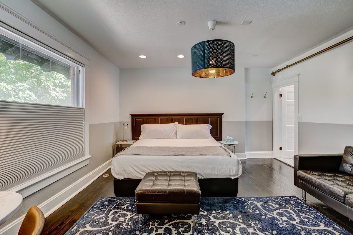 Bright bedroom with a bed, patterned rug, ottoman, couch, and large window with blinds