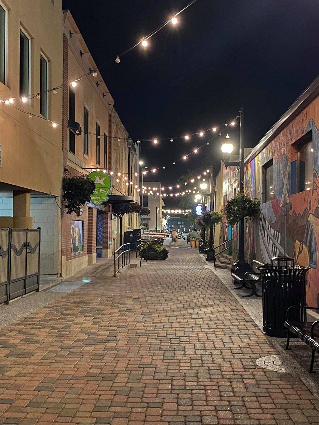 Empty brick-paved alley at night with string lights, buildings, and outdoor chairs.