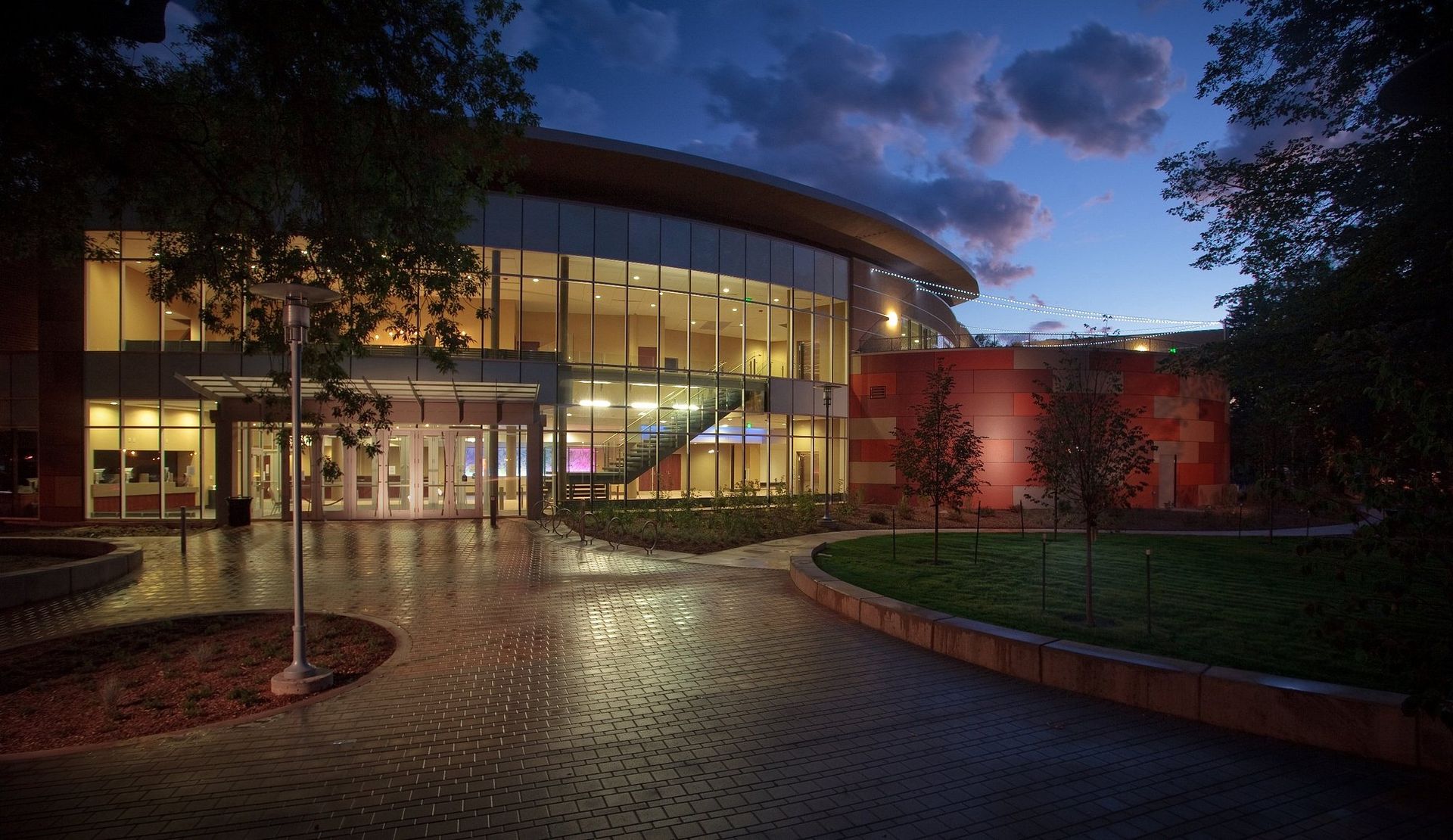 Modern glass building at dusk with lit windows, curved entrance, and wet courtyard reflecting lights