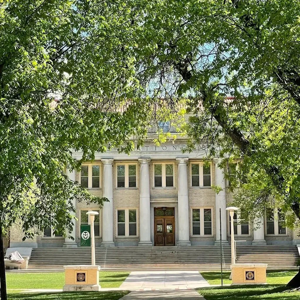 Historic stone building framed by leafy trees, with steps, columns, and a central wooden door.