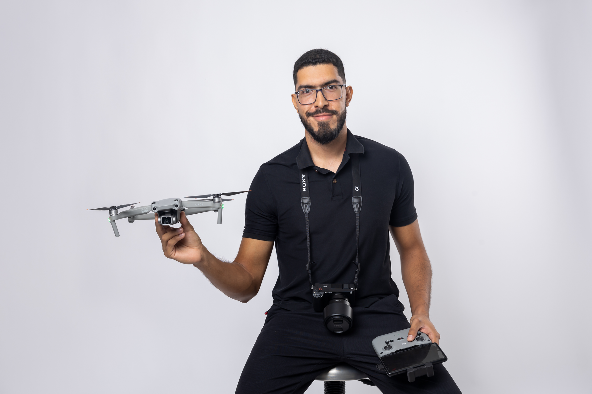 Un hombre está sentado en un taburete sosteniendo un dron y una cámara.