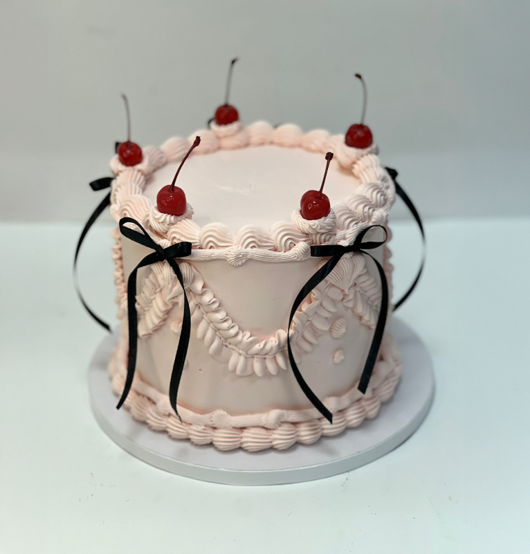 Torta quadrata glassata rosa, ciliegie in cima, decorazione con nastro nero, su un'alzata bianca.