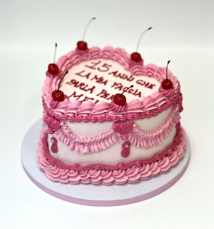 Torta rosa a forma di cuore con glassa alla crema di burro e ciliegie. Testo in italiano sulla parte superiore.
