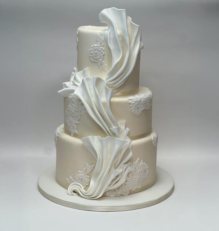 Torta nuziale bianca a tre piani con dettagli in pasta di zucchero e pizzo su base rotonda.