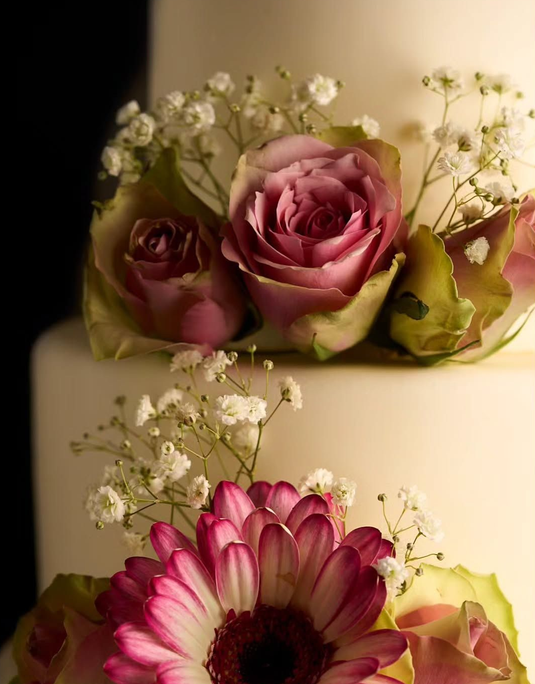 Torta nuziale decorata con rose rosa e gerbere, glassa bianca, sfondo nero.