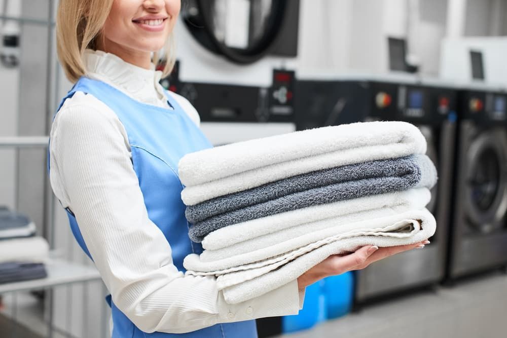 Smiling Person in a Blue Apron Holds a Neat Stack of Folded Towels — Flynns Beach Service Laundry in Port Macquarie, NSW