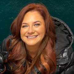Woman with red hair smiling, wearing a black jacket against a green background.