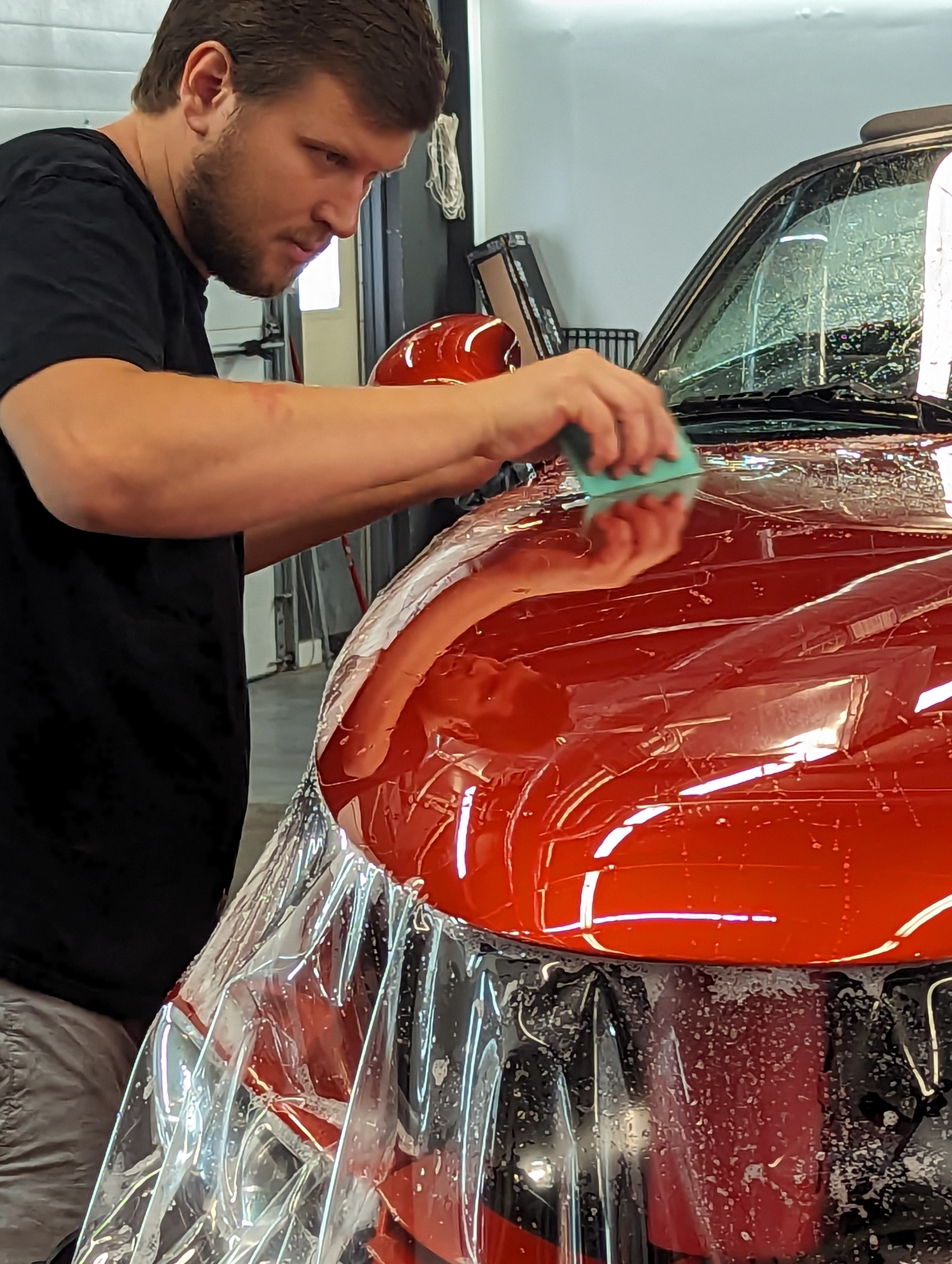 Man applying film to red car hood in a garage.