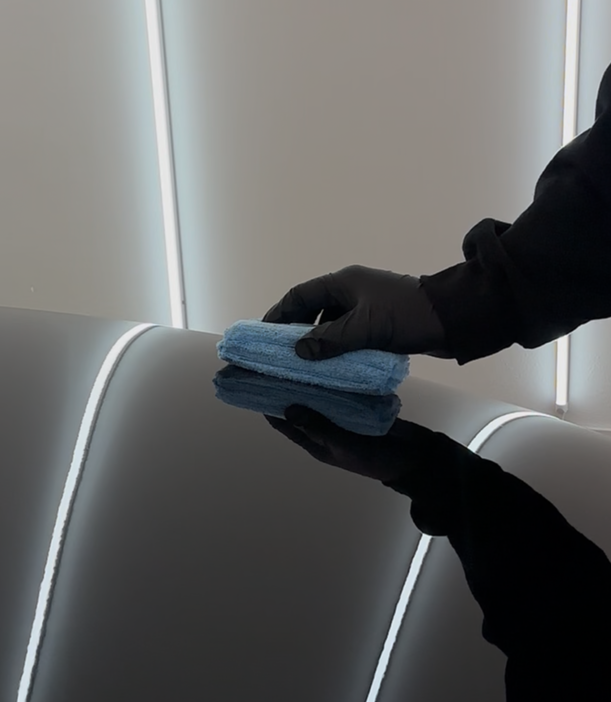 Person wearing a black glove applying car wax with a blue applicator, reflecting bright lights.