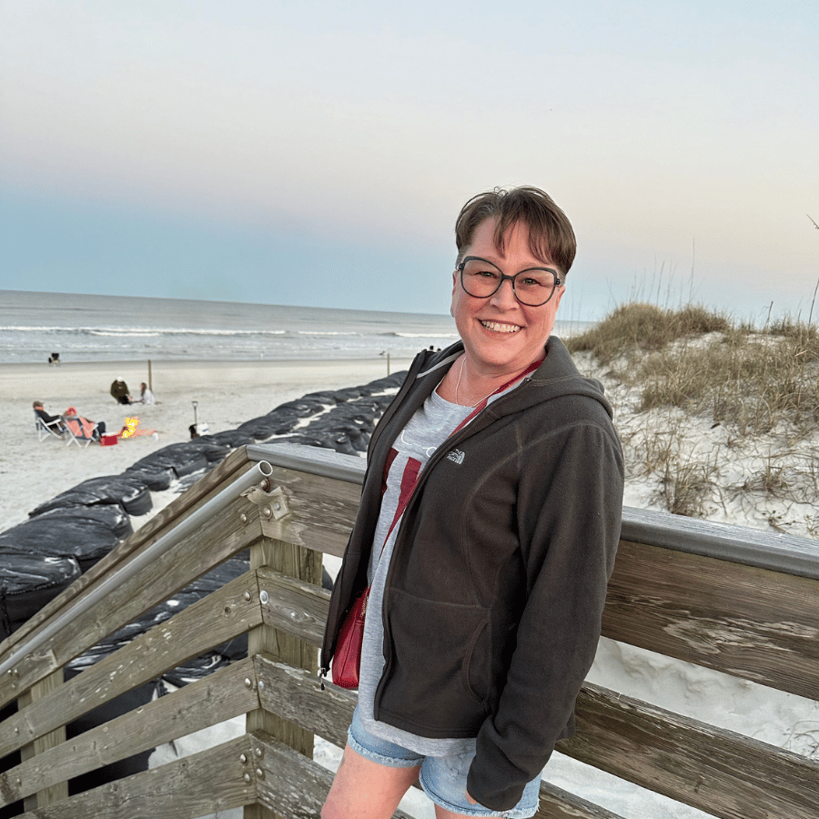 Person smiling on a wooden boardwalk by a snowy beach at sunset