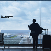 Traveler silhouetted at airport window with rolling suitcase, watching a plane take off over the runway