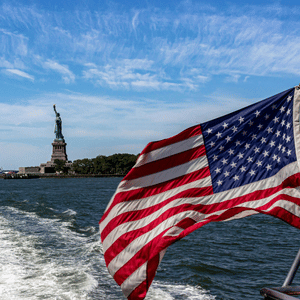 American flag waving on a boat with the Statue of Liberty in the distance over the water.