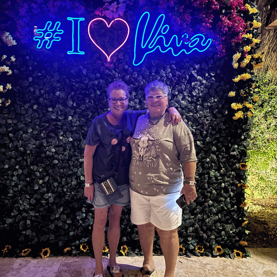 Two people posing before a neon “# I ❤️ Winx” sign on a floral wall