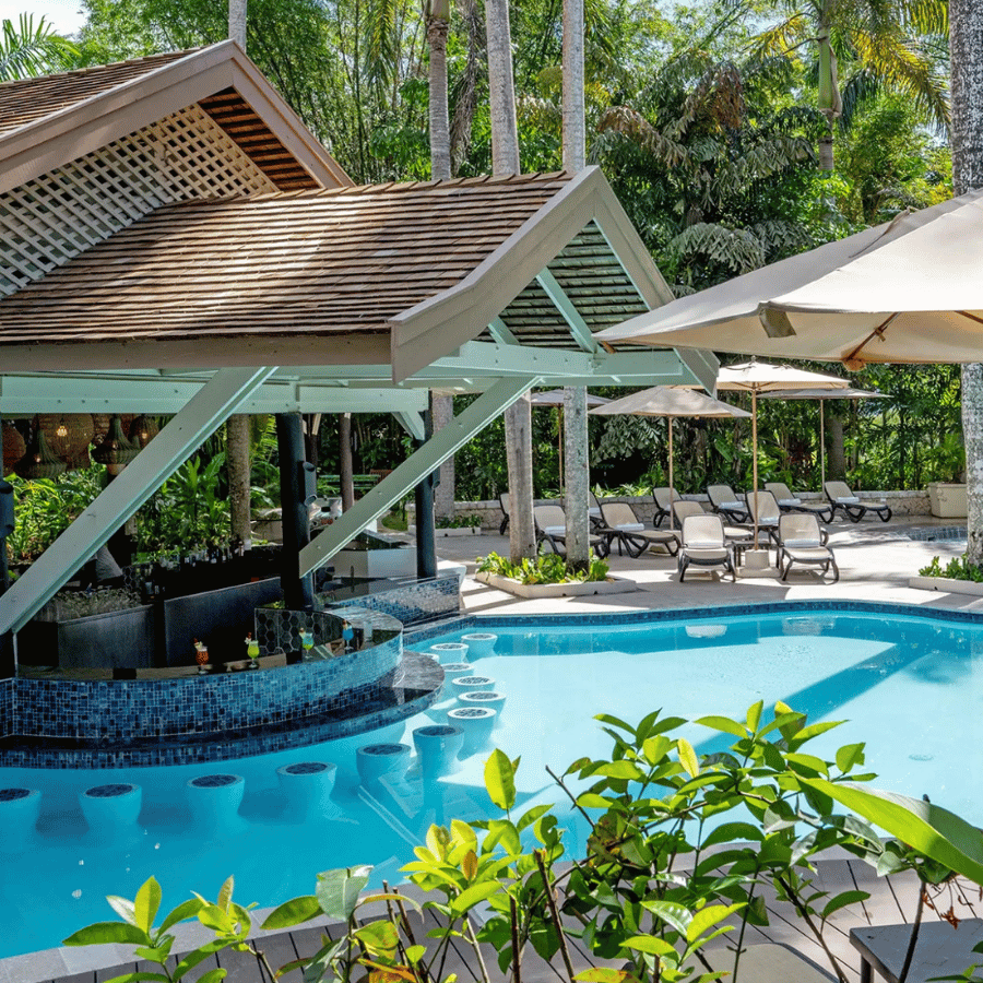 Resort pool with lounge chairs, umbrellas, and a shaded bar under tropical trees