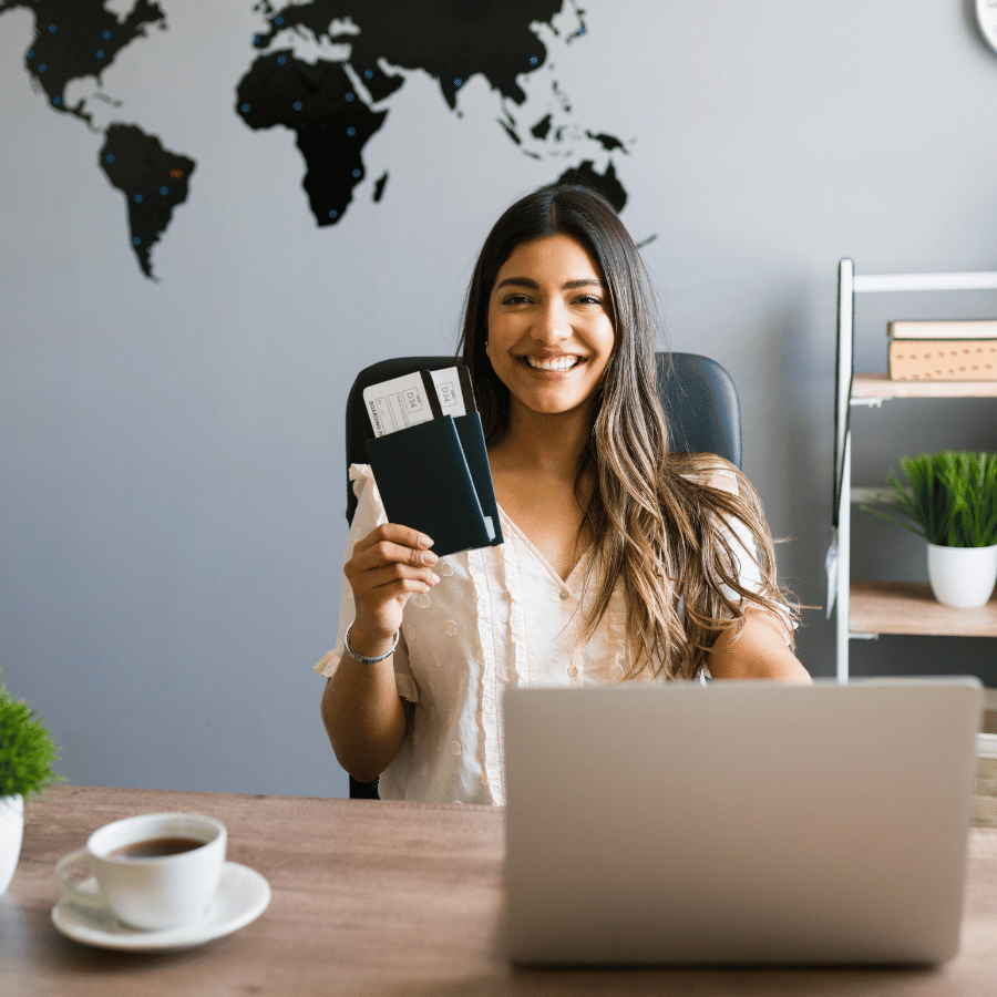 Smiling woman holds a passport at a desk with a laptop and coffee, in a home office.