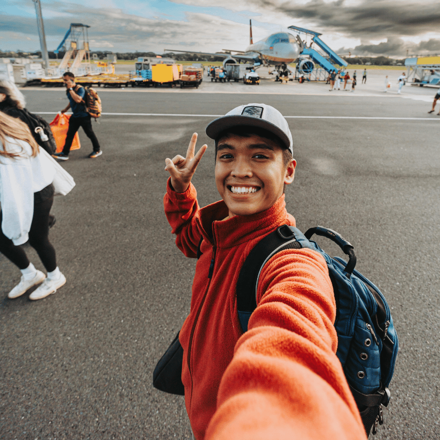 Smiling person taking a selfie on an airport tarmac, flashing a peace sign with airplanes in the background